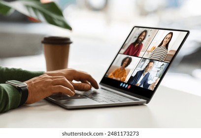 Guy sitting at a table with a laptop, using his hands to type on the keyboard. The laptop screen is displaying a video conference meeting with four participants, cafe interior