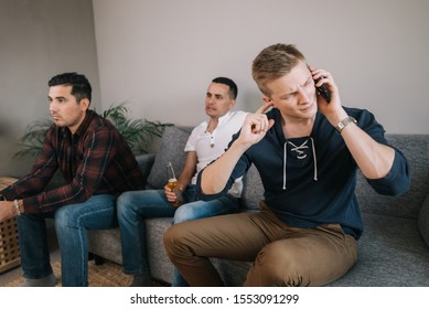 Guy Trying To Talk On The Phone. Behind The Male Back Friends Are Watching TV. Group Of Friends Watching A Football Match On TV Against The Background Of Young Man Phone Conversation.