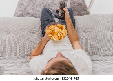 The Guy In The T-shirt Sits On The Couch, Eats Chips And Watches The TV. While Holding The Remote In His Hands.