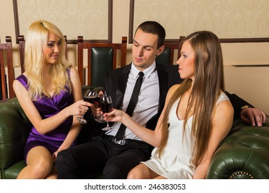 A Guy And Two Girls In The Room, Tense. Group Young People Drink Wine And Enjoy The Evening. Human Feelings - Jealousy, Love, Passion, Betrayal.