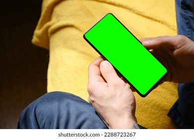 Guy Uses Green Mock-up Screen Smartphone. Young man sitting inside on yellow blanket on bed and uses phone to work or browse the Internet online, top view. Hands close-up. - Powered by Shutterstock