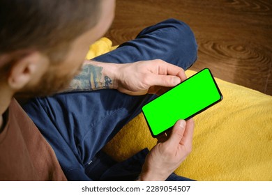 Guy Uses Green Mock-up Screen Smartphone. Young man sitting inside on yellow blanket on bed and uses phone to work or browse the Internet online, top view. Hands with multicolored tattoos close-up. - Powered by Shutterstock