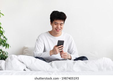 Guy Using Smartphone Browsing Internet And Texting On Phone Sits On Bed At Home. Smiling Millennial Asian Male Student In Pajamas Rest And Looks At Gadget In White Bedroom Interior, Free Space