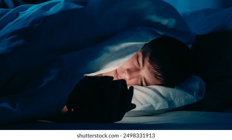 Guy using smartphone under bedsheets at night in dim light indicating addiction insomnia in bed at night. - Powered by Shutterstock