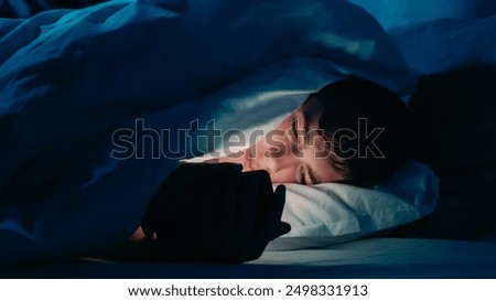 Guy using smartphone under bedsheets at night in dim light indicating addiction insomnia in bed at night.