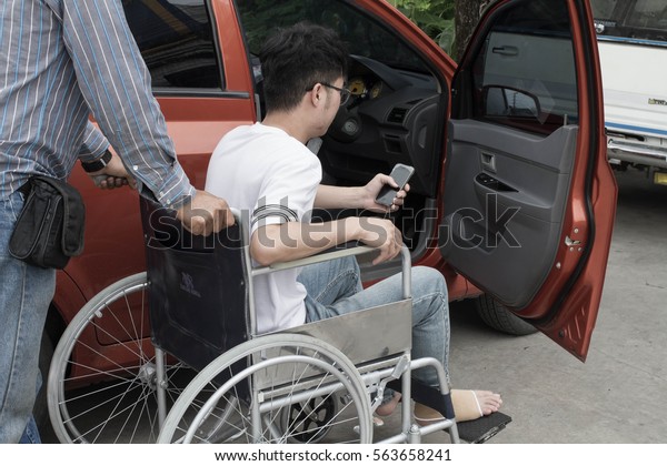 Guy wears white shirt and blue jeans sits on\
wheelchair near the door of orange car/ Father pushing wheelchair\
near son with happiness  to  clinic for treatment with team work\
-concept Help disabled\
