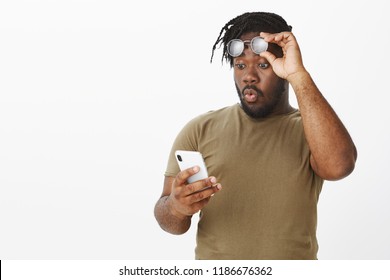 Guy whistling from surprise, being impressed with amount of money on account. Portrait of excited handsome plump african man, taking off glasses, holding rim on forehead and staring at smartphone