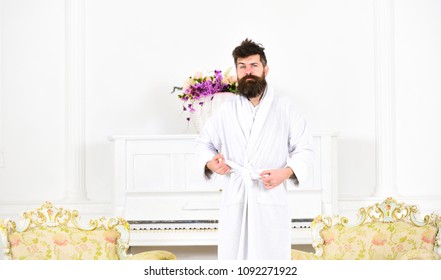 Guy In White Bathrobe Standing Between Two Antique Armchairs. Bearded Man Tightening His Robe. Sleepy Man Frowning In The Morning