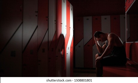 The Guy Is Worried Sitting In The Locker Room. The Athlete Is Sad In The Locker Room. Motivation And Sports. Side View Of Disappointed And Tired Sportsman Wiping Face And Leaning On Wall In Semilit