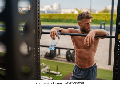 Gym enthusiast muscular man resting after a workout, leaning on a barbell rack and holding a water bottle. The outdoor gym setting includes weights and exercise equipment. - Powered by Shutterstock