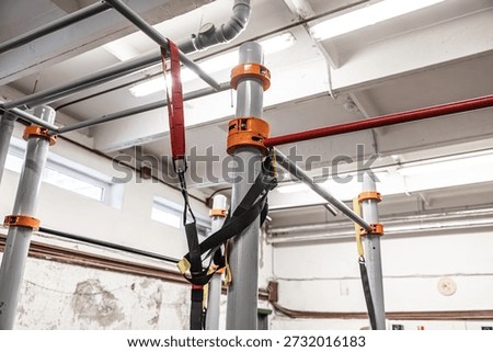 A gym monkey bar rig with gray metal poles and orange brackets, featuring attached yellow suspension training straps in a well-lit industrial workout space