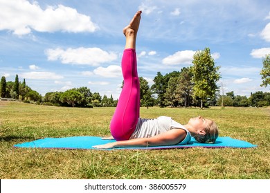 Gym And Pilates Outside - Active Young Blond Woman Exercising, Lying Down With Both Legs Up And Arms Down To Tone Them Up On Her Exercise Mat Over Sunny Blue Sky