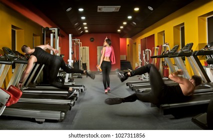  Gym With Red Walls And Blue Floor. Falling On A Treadmill. Concept: Men Fall Through A Girlfriend. They Looked At The Girl. Injury Due To Uncertainty. Injury Through Love. Beautiful Girl Charming Men