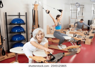 In gym, senior female performs Pilates exercises to lose weight and bring muscles of arms, chest, and body into tone using heron pulley tower. Comprehensive equipment for pumping whole body - Powered by Shutterstock