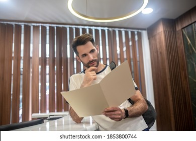 Gym Services. Male Customer Standing At Recepion Desk, Studying Carefully Price List, Touching His Chin