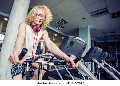 Gym. Skinny Blond Guy With Glasses And Red Headphones Trains On A Stationary Bike