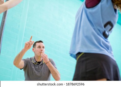 Gym Teacher With A Whistle Watching High School Students Play Netball In A Gym.