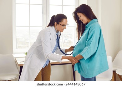 Gynecologist female doctor examining heartbeat in the abdomen of pregnant young woman with stethoscope in office in medical clinic. Smiling girl visiting her obstetrician doctor. Pregnancy check up. - Powered by Shutterstock