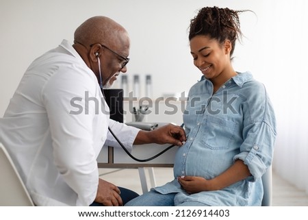 Gynecology Consultation. Smiling black pregnant woman visiting her obstetrician doctor in maternity clinic, mature male gynaecologist examining her belly with stethoscope, doing medical check-up