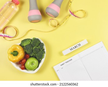 habit tracker book, wooden calendar March, gray pink dumbbells, measuring tape and vegetables in heart shape plate and bottle of water  on yellow background with copy space. Healthy food, lifestyle