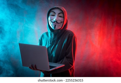 Hacker with anonymous mask with a computer and with a background of smoke and colored led