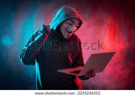 Hacker with an anonymous mask with a computer and making a fight symbol, with a background of smoke and colored leds
