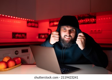 A Hacker In A Black Hoodie With A Hood On His Head Is Sitting At A Laptop Looking Straight