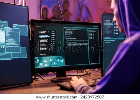 Hacker coding malicious software for password cracking in abandoned warehouse. Young man in hood programming illegal ransomware on computer with multiple monitors in hideout