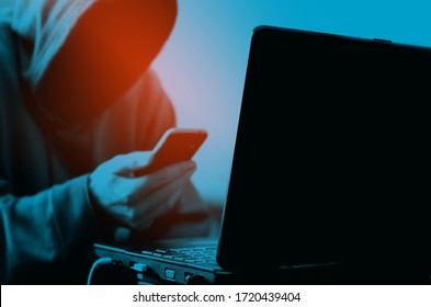Hacker. Cybercriminal. Black laptop and sitting man in hood with smartphone in hand. Blue and orange colors background. 