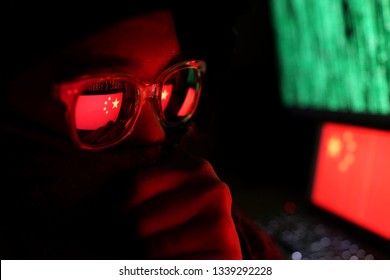 Hacker In A Dark Wearing Hoody And Cap Sitting In Front Of Computer With Digital China Flag Background And Binary Code, Ransomware Attack, Cybersecurity Concept.