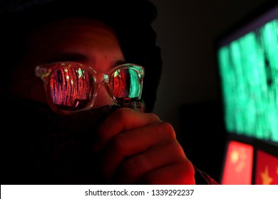 Hacker In A Dark Wearing Hoody And Cap Sitting In Front Of Computer With Digital China Flag Background And Binary Code, Cybersecurity Concept.