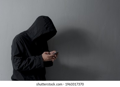 Hacker With Hidden Face In A Hoodie Uses A Mobile Phone To Hack The System Against Gray Wall