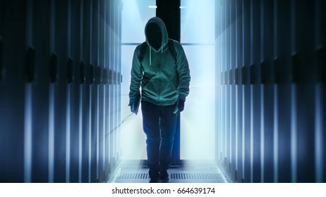 Hacker With A Hidden Face Wearing Hoodie Walks In Through Data Center Corridor.