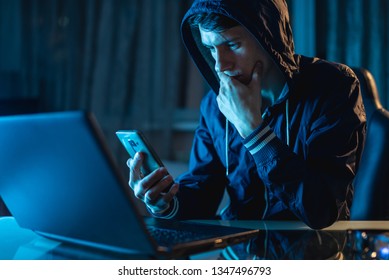 Hacker In The Hood Holding The Phone In His Hands Trying To Steal Access Databases With Passwords. The Concept Of Cyber Security