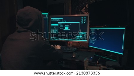 Hacker in a hood sits in front of computer screens and hacks databases. The concept of cyberterrorism, server hacking and cyberattacks. The room is dark, a person is typing code into the command line
