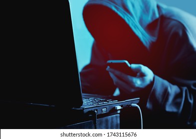 Hacker in hood without a face at laptop with phone in hand. Hacks  dark background. Internet scammer or thief. Darknet. Cyber attack.