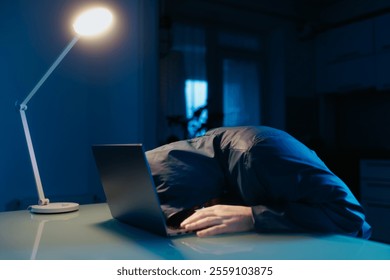 A hacker in a hoodie at a table in a dark room expresses frustration and failure, with stress evident on his face while staring at his laptop. - Powered by Shutterstock