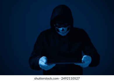 A hacker man in a black hoodie is holding a tablet in his hands. He is wearing a mask and a skull bandana. Concept of mystery and danger - Powered by Shutterstock