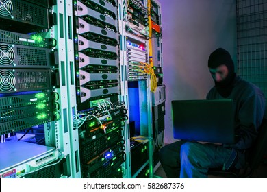 The hacker in a mask with laptop performs the attack, sitting on a chair in the server room