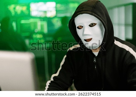 Hacker with mask programming malware using laptop in dark room with green code displayed on monitors