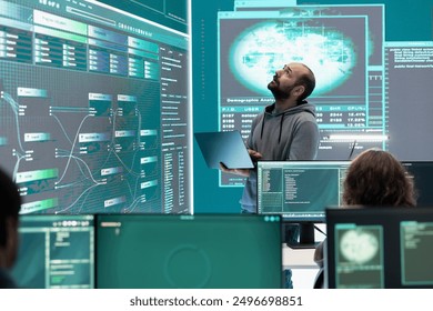 Hacker specialist working on governmental cyber surveillance in agency center, examining stolen information on a big screen during hybrid war. IT expert works on intelligence espionage. - Powered by Shutterstock