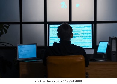 Hacker using computer at table in dark room, back view