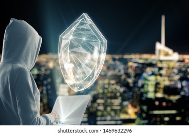 Hacker using laptop with creative glowing blue polygonal antivirus shield on blurry night city background. Safety and hacking concept. Double exposure 