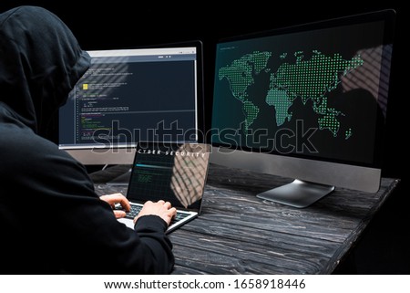 hacker using laptop with cyber security lettering on screen near computer monitors on black
