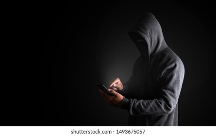 Hacker Using Smartphone. Men in black clothes with hidden face looks at smartphone screen on black background with copy space. with clipping path.