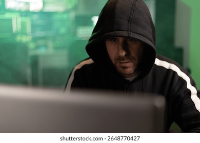 Hacker wearing a black hoodie is breaking into a secure network in a dark room with green code running on the screens