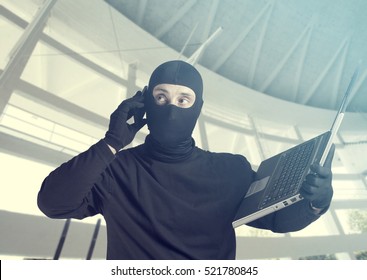 Hacker at work. Internet security concept with masked criminal holding a laptop while using a mobile phone