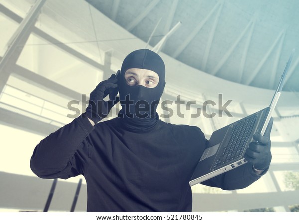 仕事中のハッカー。携帯電話を使いながら、覆面をした犯罪者がノートパソコンを持つインターネットセキュリティのコンセプト