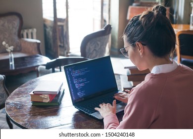 Hacking System. An Excited Girl With Dark Hair And In A Pink Sweater And Glasses Writes Code On A Laptop While Sitting In A Library. Work Undercover. Girl Hacker. View From The Shoulder.