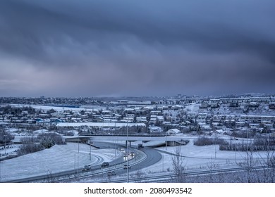 Hafnarfjordur, Iceland - November 24, 2021: A City Panorama On A Dark, Snowy Winter Day. Dark, Cloudy Sky, Residential Buildings, Cars Driving Down The Road.